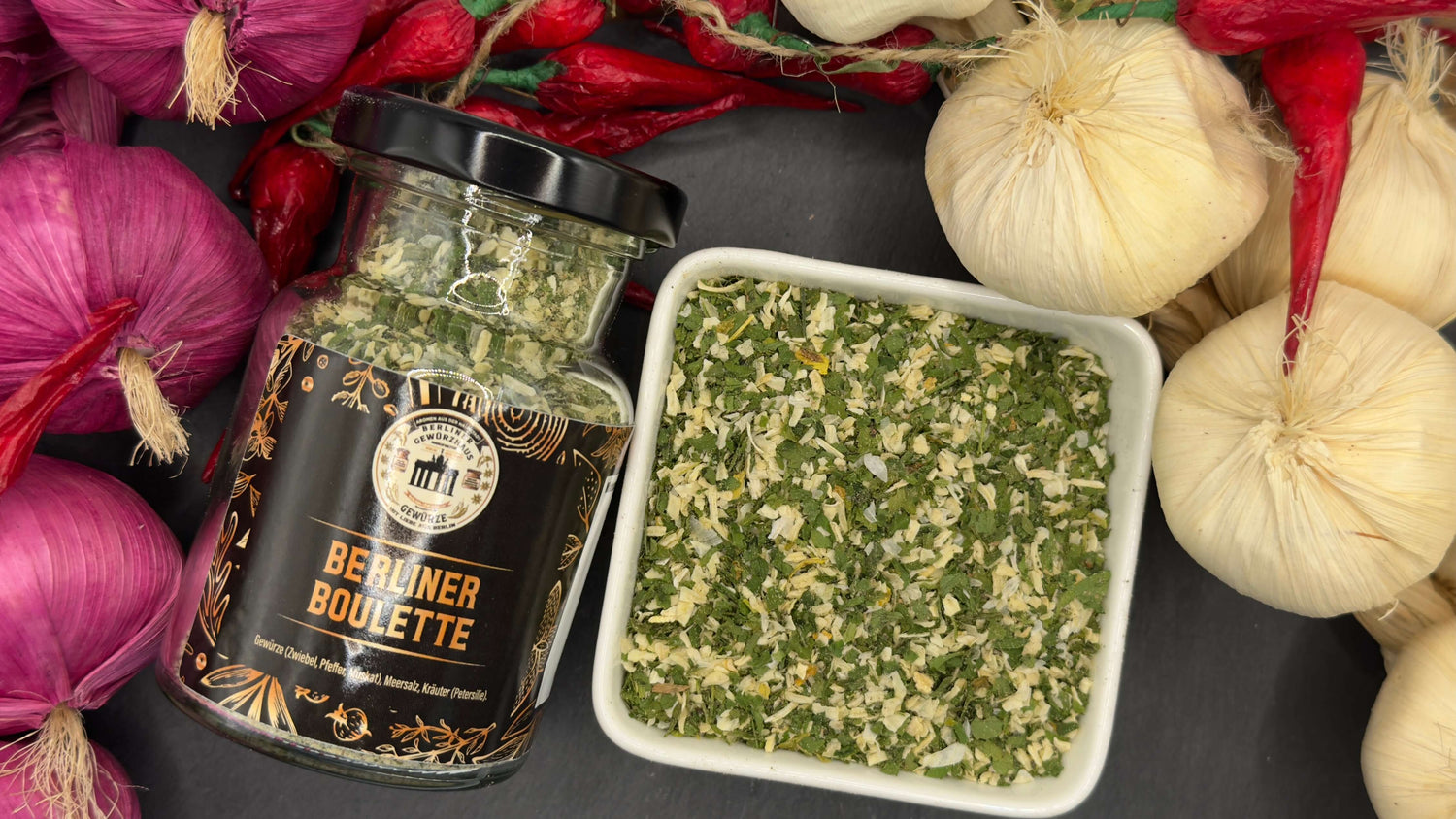 Berliner Boulette Gewürzzubereitung in einem Glas und einer Schüssel, umgeben von Zwiebeln und Knoblauch.